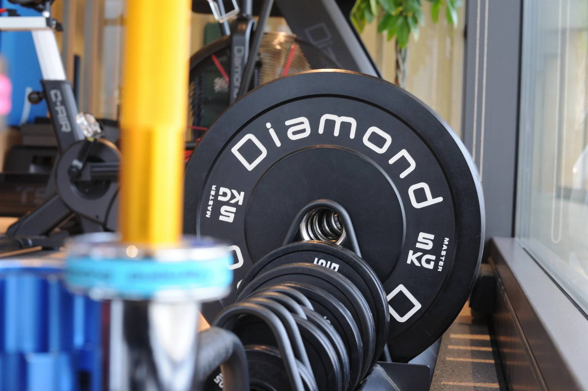 Disco bumper Diamond Master da 25 kg in primo piano su rack, con altri dischi e attrezzatura da palestra sfocata sullo sfondo.