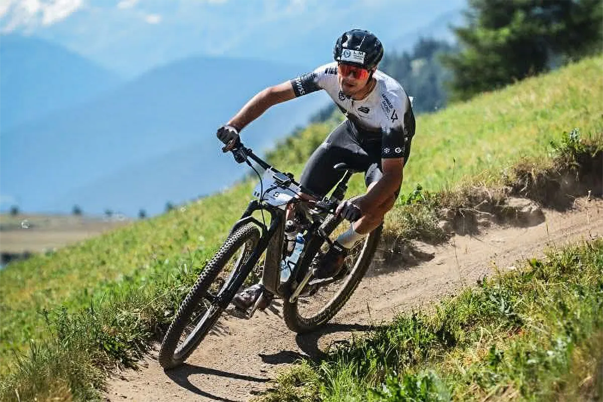 Simone Linetti in gara su mountain bike in discesa su sentiero sterrato di montagna, con occhiali rossi e maglia nera, paesaggio alpino sullo sfondo.